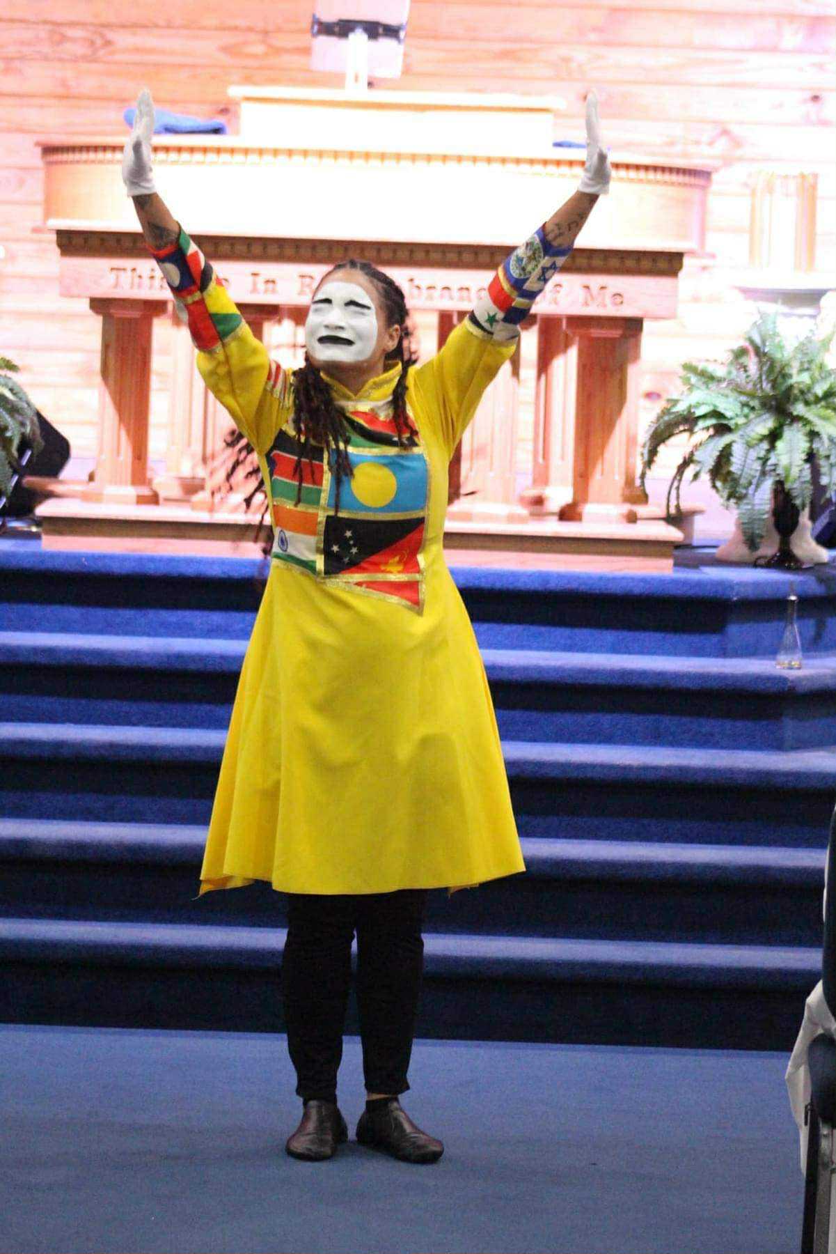 Jasmine Harris ministering through mime with raised arms, wearing a yellow garment with colorful flag panels